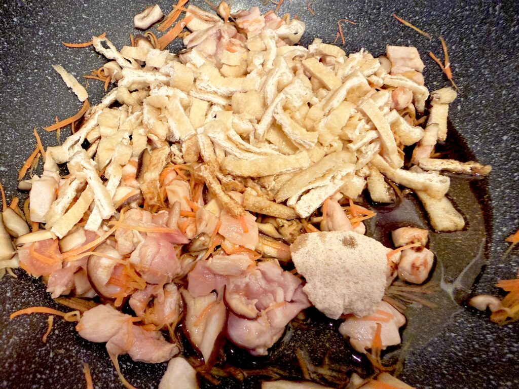 鶏肉と油揚げの混ぜご飯の具に調味料と顆粒だしを加える様子